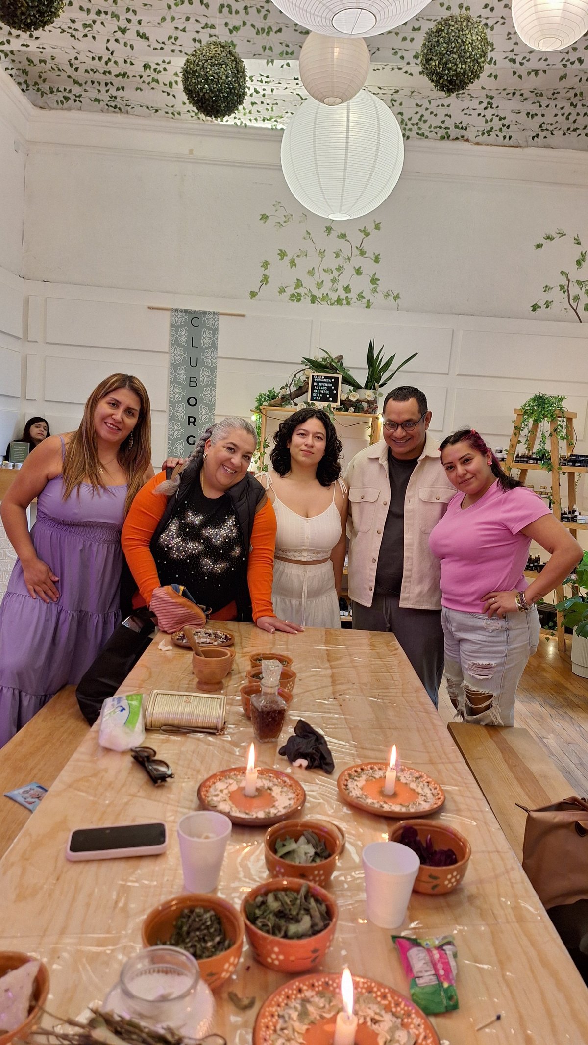 Five people posing together at a wooden table with food and plants, in a bright room decorated with hanging greenery and paper lanterns