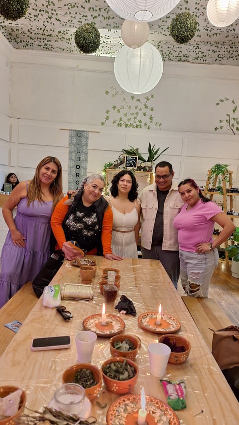 Five people stand behind a wooden table with potted plants, snacks, and decorations at a celebration in a room with hanging greenery and white paper lanterns